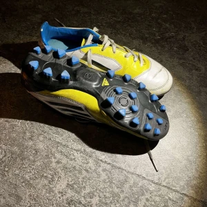 Adidas fotbollsskor i gult och vitt - Säljer ett par Adidas fotbollsskor i gult och vitt med blå detaljer. Skorna har snörning och dobbar för bra grepp på planen. Perfekta för fotbollsträning och matcher.
