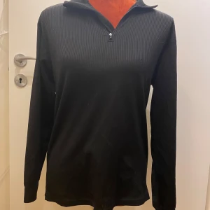 Underställ tröja Pouge M Svart - Snygg svart ribbad tröja från Pogue med dragkedja vid halsen. Perfekt för en stilren look. Tröjan har långa ärmar och en bekväm passform. underställ med dragkedja i halsen Längd: 67 cm Armhåla till armhåla: 46 cm   Inga bilder när den sitter på mig! Katter finns i hemmet