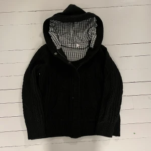 Svart stickad hoodie med rutigt foder - Säljer en svart stickad hoodie med ett snyggt rutigt foder i huvan. Tröjan har knappar framtill och långa ärmar, perfekt för kyliga dagar. Den är både stilren och bekväm.