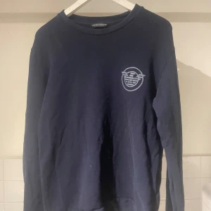 Mörkblå tröja från Armani - Säljer en mörkblå tröja från Armani med ett tryck på bröstet. Tröjan har långa ärmar och en rund halsringning. Perfekt för en avslappnad stil.