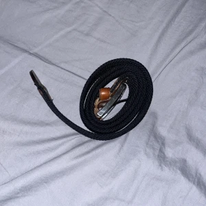 Svart flätat bälte - Ett skönt Snyggt svart flätat bälte som också är stretchat med detaljer i brunt läder. Perfekt för att ge en stilren touch till din outfit. Bältet har en klassisk design med en silverfärgad spänne.