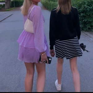 Syrenlila klänning - Charmig lila klänning med prickigt mönster och volanger. Den har en omlott-design med knytband i midjan och långa ärmar. Perfekt för sommarens festligheter.