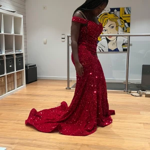 Röd glittrig långklänning - Fantastisk röd glittrig långklänning med offshoulder-design. Klänningen har en elegant passform och ett släp som ger en dramatisk effekt. Perfekt för speciella tillfällen där du vill glänsa.