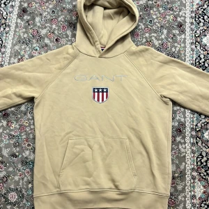 GANT tröja - Säljer en snygg beige hoodie från GANT med ett broderat emblem på bröstet. Hoodien har en klassisk känguruficka och en bekväm huva. Perfekt för en avslappnad stil. Storlek 176 men passar Xs,S