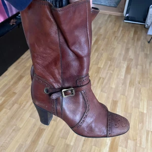 Bruna skinnstövletter med klack - Snygga bruna stövletter i skinn med dekorativt bälte och klack. Perfekta för höst och vinter med en elegant spetsig tå och fina detaljer.