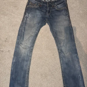 Rock revival jeans - Snygga blå jeans med unika broderade detaljer på bakfickorna. Jeansen har en straight passform och är tillverkade i denim. Perfekta för dig som vill ha en stilren men ändå iögonfallande look.