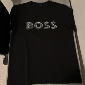 Svart t-shirt från Boss - Snygg svart t-shirt från Boss med ett stort, stilrent tryck av märkesnamnet på framsidan. Perfekt för en casual look med en touch av lyx. Kortärmad och i en klassisk passform. Helt oanvänd fick hem den och den var för stor.