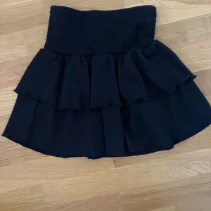 Svart volangkjol - Snygg svart kortkjol med volanger. Perfekt för att ge en trendig touch till din outfit. Den elastiska midjan gör den bekväm och lätt att bära. Lappen är avklippt men passar mig som har xs