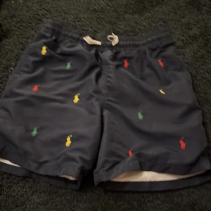 Svarta bad shorts från Polo Ralph Lauren - Snygga svarta shorts från Polo Ralph Lauren med färgglada broderade logotyper i rött, grönt och gult. De har en bekväm passform med snörning i midjan. Perfekta för en avslappnad stil.
