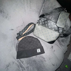 Grå axelväska med monogram. Svart mössa med monogram. Svart skärp med monogram  - Snygg grå axelväska från Louis Vuitton med klassiskt monogrammönster. Väskan har flera fack med dragkedja och en justerbar axelrem i svart och grått rutmönster. Perfekt för att bära dina nödvändigheter med stil.