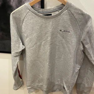 Grå sweatshirt från Bondelid - Säljer en stilren grå sweatshirt från Bondelid med broderad logga på bröstet. Tröjan har långa ärmar med röd och vit randig detalj längs ärmarna. Perfekt för en avslappnad stil.