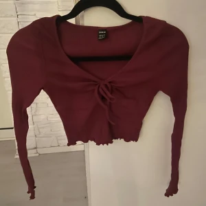 Vinröd ribbad topp från Shein - Säljer en vinröd ribbad topp från Shein med knytning framtill. Toppen har långa ärmar och en croppad design som ger en trendig look. Perfekt för att styla med höga jeans eller kjol.
