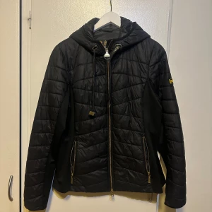 Svart quiltad jacka från Barbour - Säljer en stilren svart quiltad jacka från Barbour International. Jackan har en praktisk huva och dragkedja framtill. Den är perfekt för kyligare dagar och har en klassisk design med Barbour-logga på ärmen. 