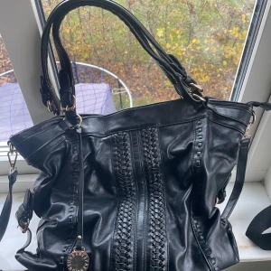 Svart axelväska med flätade detaljer - Snygg stor svart axelväska med unika flätade detaljer på framsidan. Väskan har dubbla handtag och en avtagbar axelrem för extra flexibilitet. Perfekt för att bära med stil.