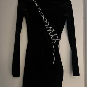 Svart klänning med snörning - Elegant svart klänning i sammet med långärmad design och unik snörning över bröstet. Perfekt för en stilfull kväll.