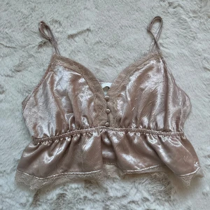 Beige glansig topp från H&M - Säljer en elegant beige glansig topp från H&M med spetsdetaljer och knappar framtill. Toppen har smala axelband och en v-ringad design. Perfekt för en stilfull look.