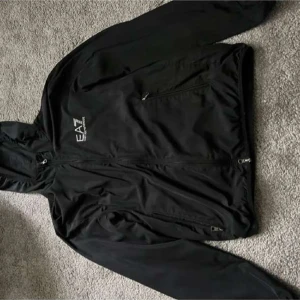 Svart jacka med huva från EA7 - Snygg svart jacka från EA7 med huva och dragkedja framtill. Perfekt för en sportig look. Jackan har en stilren design med logotypdetalj på bröstet. Har använt två gånger bara 