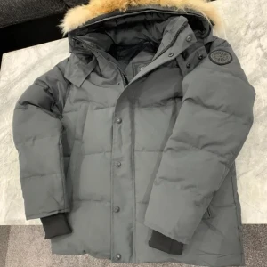 Grå Canada goose black label - Snygg och varm grå dunjacka med pälsdetalj på huvan. Jackan har en dragkedja och knappar framtill samt ribbade ärmslut för extra komfort. Perfekt för kalla vinterdagar.