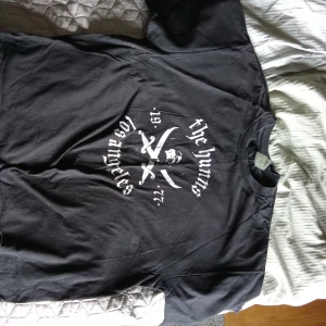 Svart t-shirt med tryck - Säljer en svart t-shirt med olika tryck. Den första har texten 'The Huns Los Angeles' med ett dödskallemotiv. Den andra har texten 'U.S. BOMBS' med stjärnor. Den tredje har ett motiv med texten 'Duane Peters' och en belöningstext. Perfekt för dig som gillar unika och tuffa tryck!