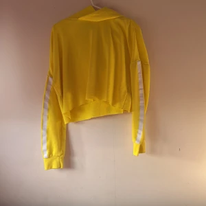 Gul croppad hoodie - Säljer en gul croppad hoodie med långa ärmar och vita ränder längs ärmarna. Perfekt för en sportig look eller avslappnade dagar.