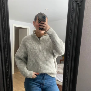 Grå stickad tröja med dragkedja från Sarah Hester - Mysig grå stickad tröja från carin Wester med en praktisk dragkedja vid halsen. Tröjan har en avslappnad passform och långa ärmar, perfekt för kyliga dagar. Den ribbade strukturen ger en stilren look.