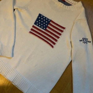 Beige stickad tröja från Gant - Snygg beige stickad tröja från Gant med ett broderat amerikanskt flaggmotiv på framsidan. Tröjan har långa ärmar och ribbade muddar. På ärmen finns texten 'Gant New Haven 1949' broderad i blått.