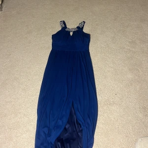 Blå långklänning från Speechless - Elegant blå långklänning från Speechless med glittriga detaljer vid halsringningen. Klänningen har en vacker fall och är ärmlös, perfekt för speciella tillfällen. Tillverkad i ett mjukt och flödande material som ger en lyxig känsla.