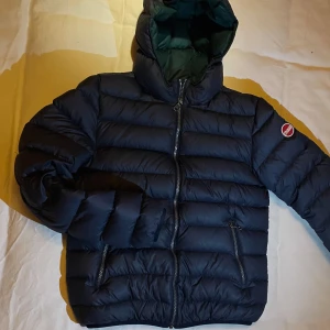 Mörkblå dunjacka med huva - Blå Colmar dunjacka med huva och dragkedja, grön insida.  Jackan har en vadderad design och en liten logotyp på ärmen. Perfekt för kyliga dagar. Observera den lilla lagningen på bilden, syns knappt alls om man inte vet om den. 