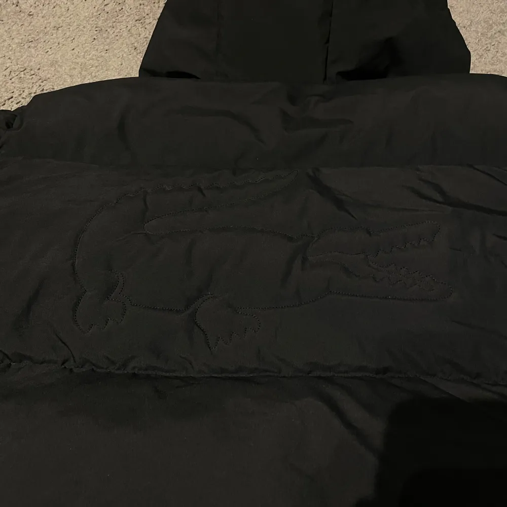 Snygg svart dunjacka från Lacoste med huva och dragkedja. Jackan har en diskret broderad logotyp på bröstet och en stor disorder logga på ryggen. Den är perfekt för kyligare väder. Den har en klassisk design med praktiska fickor och justerbara snören vid fållen.. Takit.