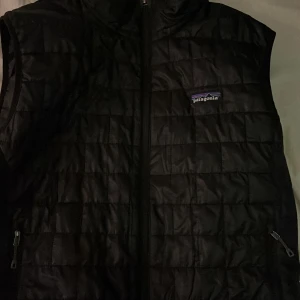 Svart dunväst från Patagonia - Säljer en stilren svart dunväst från Patagonia. Västen har en dragkedja framtill och är quiltad för extra värme. Perfekt för kyliga dagar och passar bra över en tröja eller skjorta.