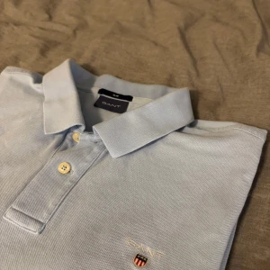 Ljusblå pikétröja från GANT - Säljer en stilren ljusblå pikétröja från GANT med klassisk krage och knappar framtill. Tröjan har en broderad logotyp på bröstet och är perfekt för en avslappnad men snygg look.