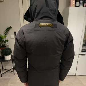 Svart dunjacka från Canada Goose - Säljer en svart dunjacka från Canada Goose med huva och knappar framtill. Jackan har en broderad logotyp på ärmen och är perfekt för kalla vinterdagar. Den har en stilren design med praktiska fickor.