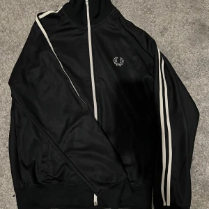 Svart track jacket från Fred Perry - Snygg svart track jacket från Fred Perry med vita ränder längs ärmarna och broderad logga på bröstet. Jackan har dragkedja och är i nyskick. Passar nog dig som är mellan 170-180 centimeter. Tveka inte på att höra av er vid några som helst funderingar.