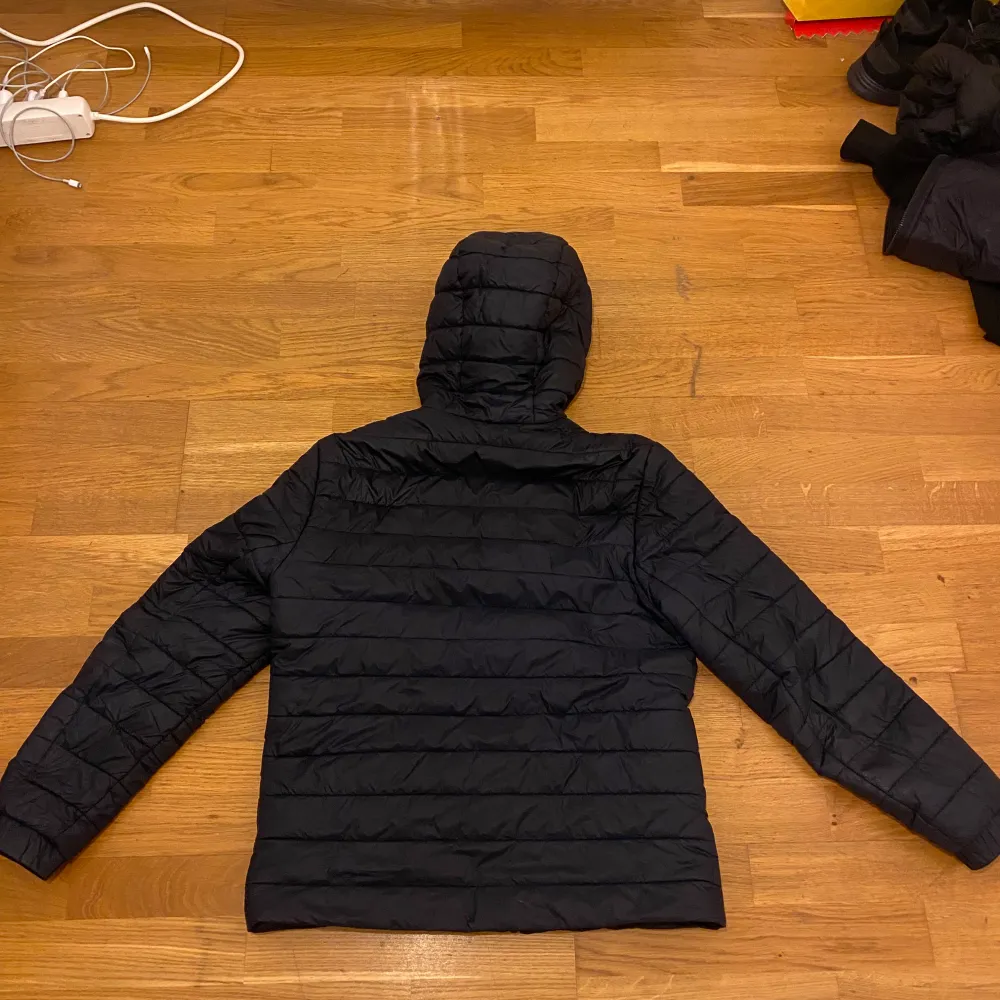 Snygg svart quiltad jacka från Hugo med huva och dragkedja. Perfekt för kyligare dagar. Jackan har en stilren design med märkets logga i rött på bröstet. Och använd knappt men de finns bara ett litet hål där ni ser och bakom bara en jätte liten scratch . Takit.
