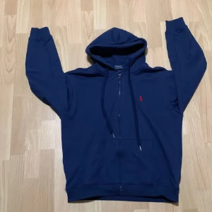 Mörkblå hoodie från Polo Ralph Lauren - Mörkblå hoodie från Polo Ralph Lauren med dragkedja och huva, strl L men passar M/L. Röd logga på bröstet och justerbara snören i huvan. Använd ett fåtal gånger och i nyskick. 