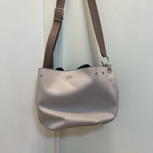 axelväska/handväska  från Guess - Snygg beige axelväska från Guess med justerbar rem och stilren design. Väskan har en praktisk storlek och är perfekt för att bära dina dagliga nödvändigheter.Har två band så går att ha som både handväska och axelväska .Den har en magnetisk stängning och flera fack inuti för enkel organisering. Den är slutsåld,DEN ÄR ÄKTA! Pris går att diskuteras så tveka inte att höra av dig!🥰🎄