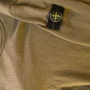 Olivgrön tröja från Stone Island - Snygg olivgrön tröja från Stone Island med deras ikoniska märke på ärmen. Perfekt för en stilren och avslappnad look.