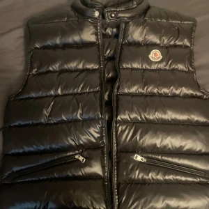 Svart dunväst från Moncler - Säljer en stilren svart dunväst från Moncler med glansig yta och dragkedja.  Västen är i storlek 4 som ungefär motsvarar M. 