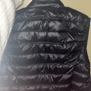 Svart dunväst från Emporio Armani - Snygg svart dunväst från Emporio Armani med dragkedja framtill. Perfekt för kyligare dagar och ger en stilren look. Märkeslogga synlig på bröstet.