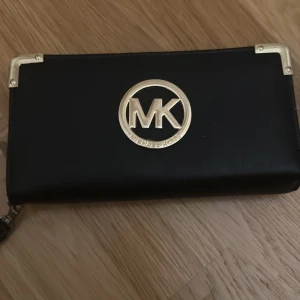Svart plånbok från Michael Kors - Elegant svart plånbok från Michael Kors med guldfärgade detaljer och logotyp på framsidan. Plånboken har flera fack för kort och en dragkedja i mitten för mynt. Perfekt för att hålla ordning på dina tillhörigheter med stil. Pris kan diskuteras. 
