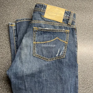Blå jeans från Jacob Cohen - Säljer ett par snygga blå jeans från Jacob Cohen. De är handgjorda med utvalda japanska tyger och italienskt hantverk. Jeansen har en klassisk femficksdesign och knappar i gylfen. Perfekta för dig som vill ha stil och kvalitet.