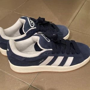 Blå Adidas Campus sneakers - Snygga blå Adidas Campus sneakers med vita detaljer och klassiska tre ränder på sidan. Skorna är oanvända och perfekt skick.