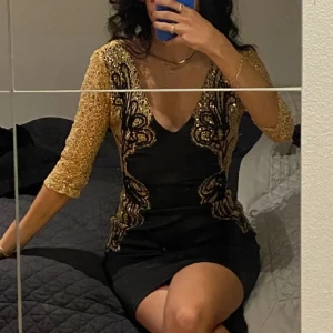 Svart och guld klänning med paljetter (nyårsklänning) - Snygg svart klänning med guldpaljetter och v-ringad design. Klänningen har trekvartsärmar och ett detaljerat mönster i guld som ger en elegant touch. Perfekt för en festlig kväll!