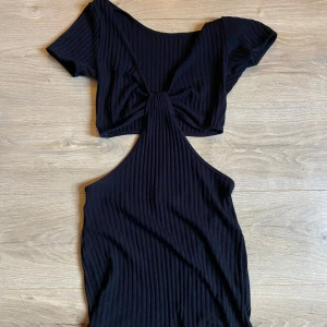 Svart ribbad klänning med öppen rygg - Trendig svart ribbad klänning med korta ärmar och en snygg öppen rygg. Klänningen har en knytning framtill och en kort längd som ger en modern look.