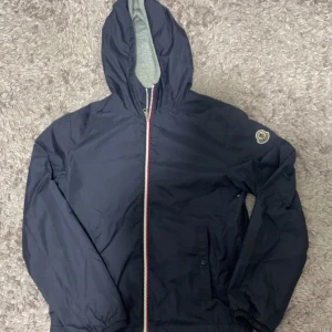 Mörkblå jacka från Moncler - Snygg mörkblå jacka från Moncler med huva och dragkedja i rött, vitt och blått. Jackan har ett broderat Moncler-märke på ärmen och är perfekt för kyligare dagar. Jackan är 100% äkta och 800 är ett skitbra pris jackan är storlek xs/s 