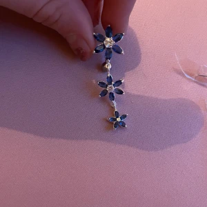 Silver navelpiercing - Snygga navelpiercingar med blå blommor och silverdetaljer. De har en böjd stav med en kula i ena änden och två hängande blommor i olika storlekar. Perfekt för att ge en extra touch till din stil.