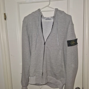Grå zip hoodie Stone Island - Säljer en grå huvtröja från Stone Island med dragkedja och ikonisk logga på ärmen. Tröjan har en bekväm passform och är perfekt för en avslappnad stil. Den har långa ärmar och en praktisk huva.