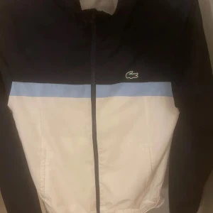 Svart och vit hoodie från Lacoste - Snygg och sportig hoodie från Lacoste i svart och vitt med en ljusblå rand. Den har en dragkedja framtill och det klassiska Lacoste-märket på bröstet. Perfekt för en avslappnad stil.