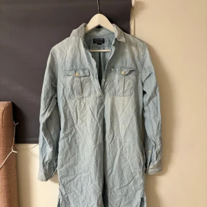 Ljusblå jeansklänning från Polo Jeans Company - Snygg ljusblå jeansklänning från Polo Jeans Company med klassisk krage och knappar framtill. Klänningen har långa ärmar och två bröstfickor med knappdetaljer. Perfekt för en avslappnad stil.