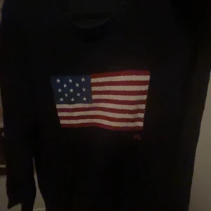 Polo Ralph Lauren, The Iconic Flag Jumper - Snygg svart tröja från Ralph Lauren med en stor amerikansk flagga på framsidan. Perfekt för en casual look med en touch av patriotism. Tröjan har långa ärmar och en klassisk passform. Säljs för 6 095kr hos Ralph Laurens hemsida. Storlek L